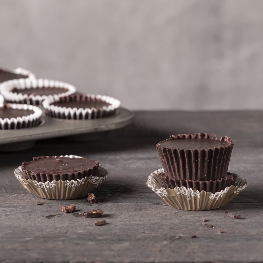 Two stacked chocolate cups in paper liners, one with a bite taken out, sit on a dark surface. Several more chocolate cups are in a muffin tin in the background. Chocolate crumbs are nearby.