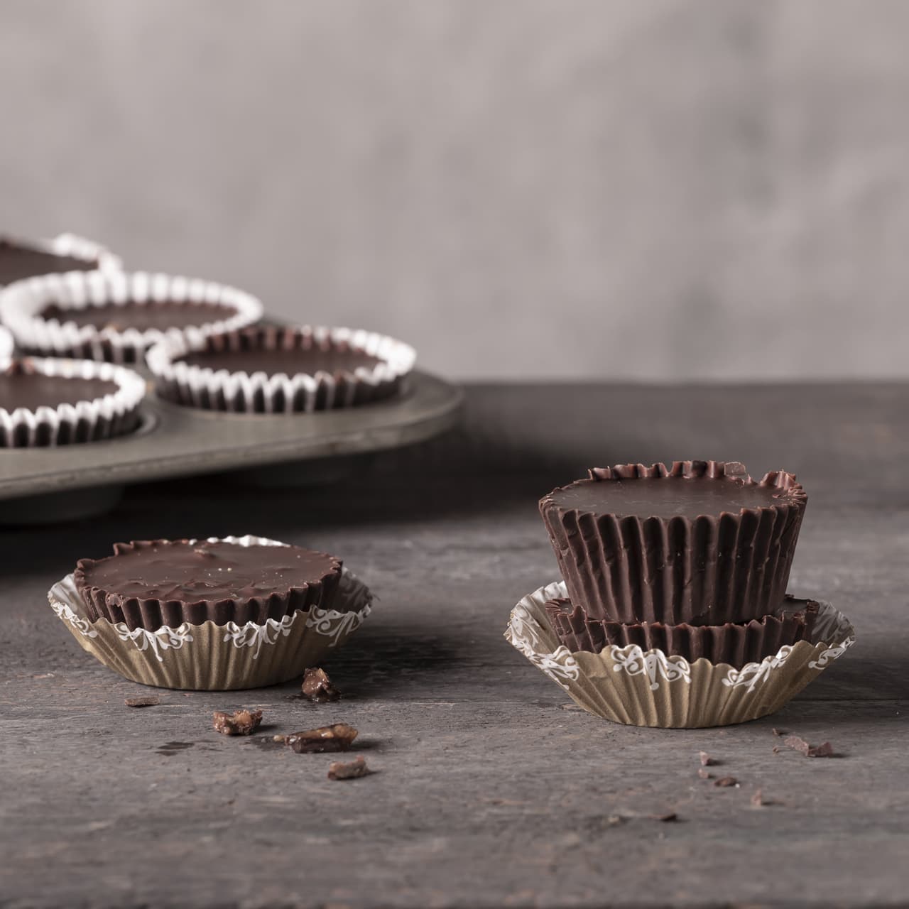 Two stacked chocolate cups in paper liners, one with a bite taken out, sit on a dark surface. Several more chocolate cups are in a muffin tin in the background. Chocolate crumbs are nearby.