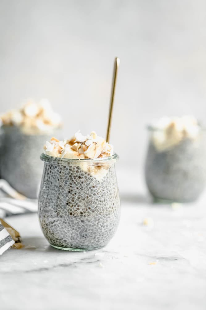 A small glass jar filled with chia pudding, topped with whipped cream and toasted coconut, with a spoon inside; two similar jars are in the background.