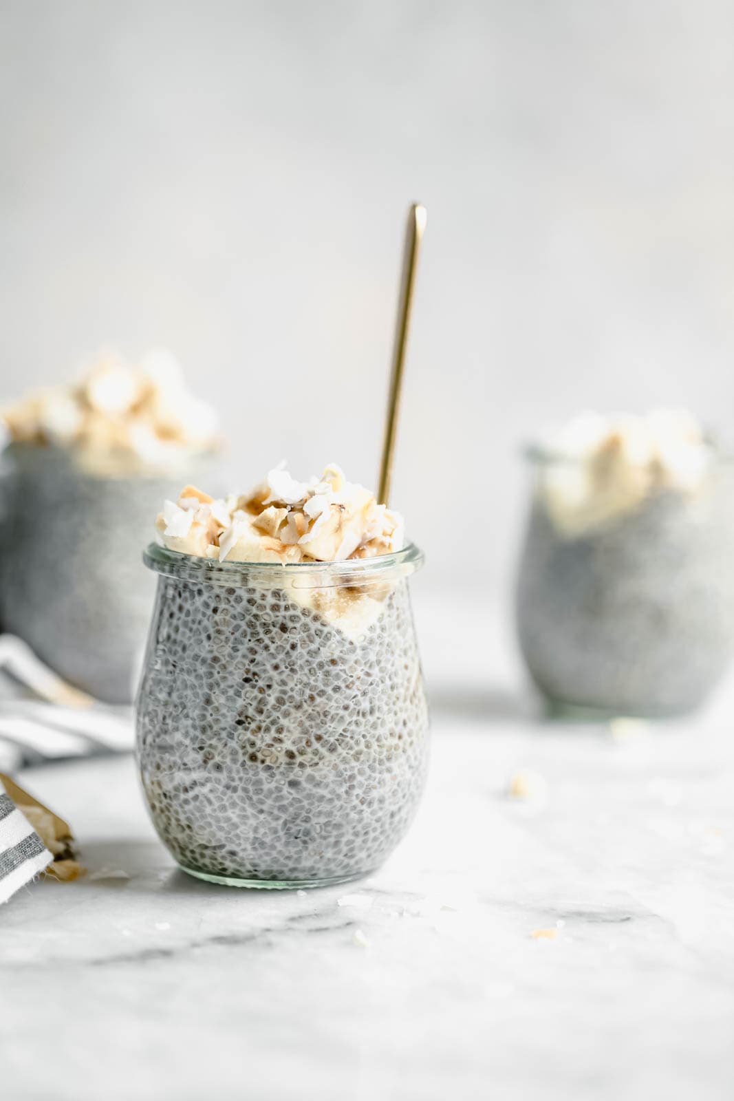 A small glass jar filled with chia pudding, topped with whipped cream and toasted coconut, with a spoon inside; two similar jars are in the background.