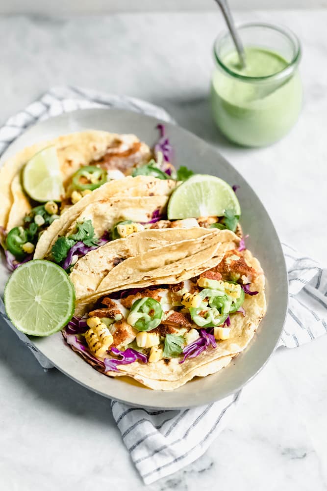 A plate with three tacos filled with grilled corn, jalapeño, cabbage, cilantro, and lime wedges, next to a jar of green sauce with a spoon.