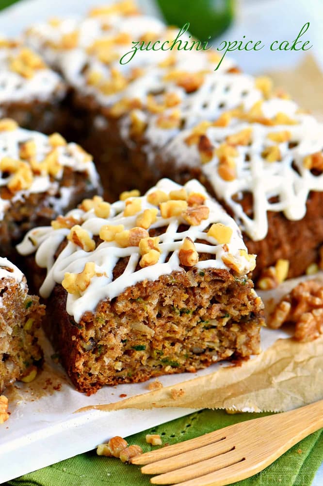 Sliced zucchini spice cake topped with white icing and chopped walnuts, displayed on parchment paper with a wooden fork nearby.