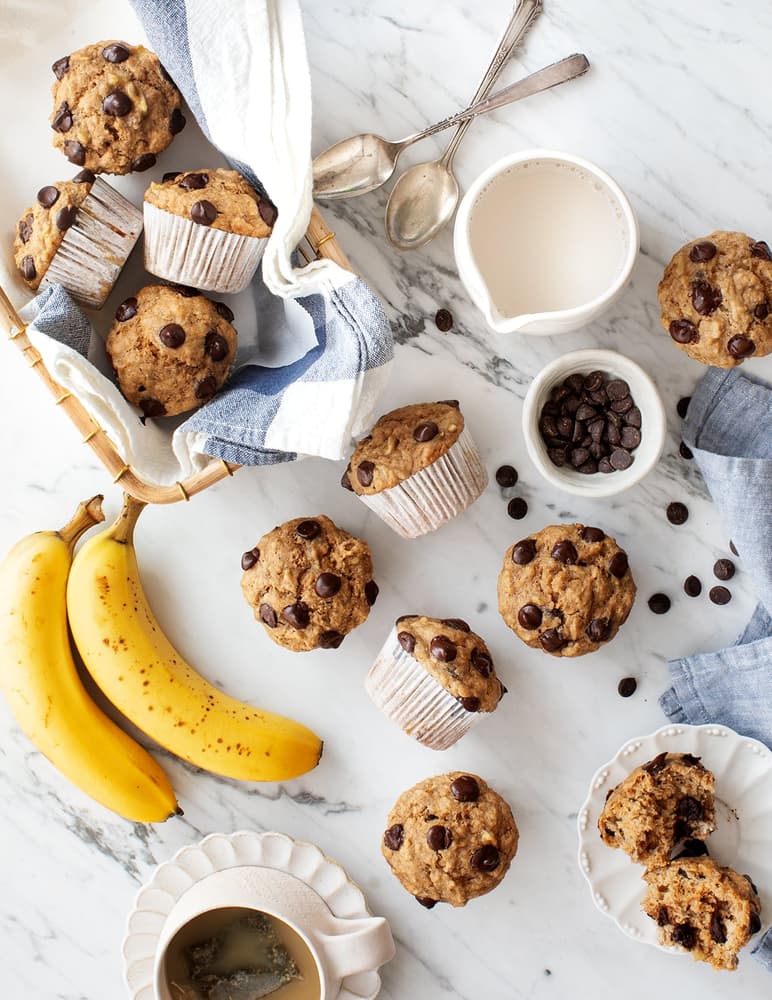 Banana chocolate chip muffins on a marble surface with fresh bananas, chocolate chips, a cup of coffee, a jug of milk, and spoons; some muffins are unwrapped or cut open.