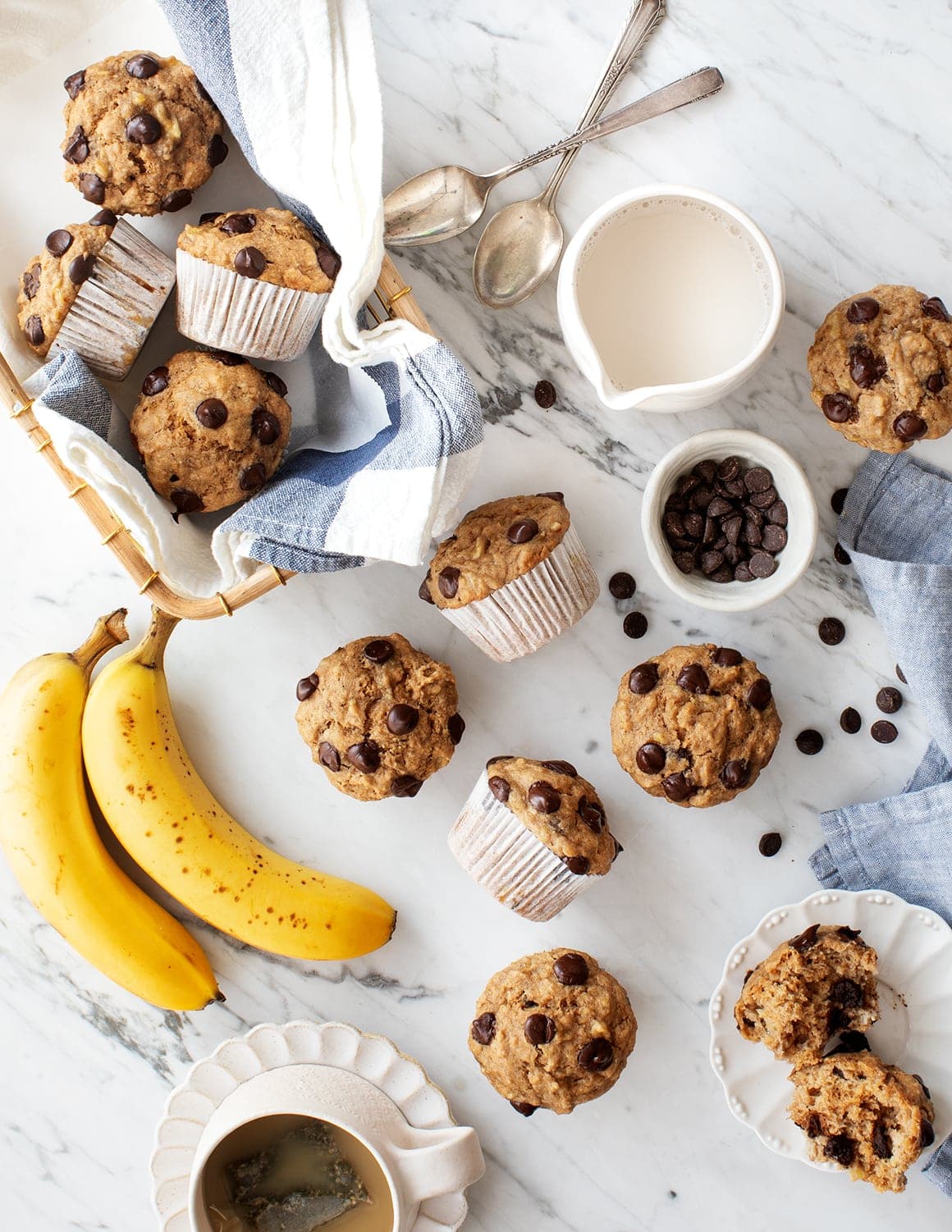 Banana chocolate chip muffins on a marble surface with fresh bananas, chocolate chips, a cup of coffee, a jug of milk, and spoons; some muffins are unwrapped or cut open.