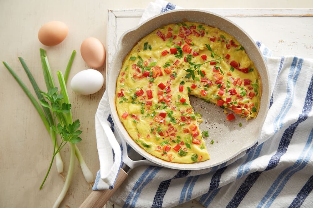A frittata with diced red peppers and green onions in a pan, with fresh eggs, parsley, and green onions beside it on a striped cloth.