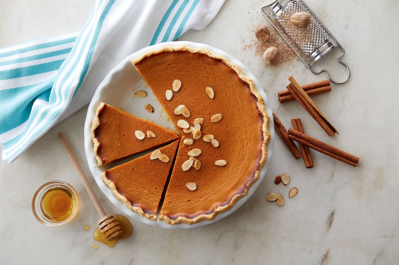 Pumpkin pie with two slices missing, topped with almond slivers. Surrounding items include a honey dipper, honey jar, cinnamon sticks, whole nutmeg, grater, and a striped towel.