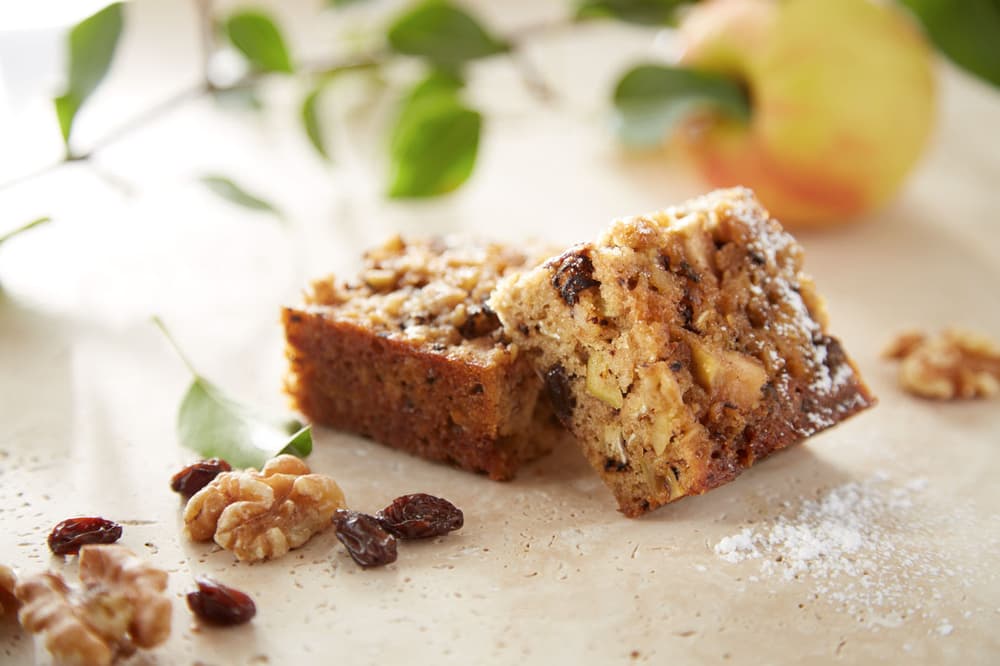 Two pieces of nut and fruit cake sit on a light surface, surrounded by walnuts, raisins, powdered sugar, and a blurred apple and leaves in the background.