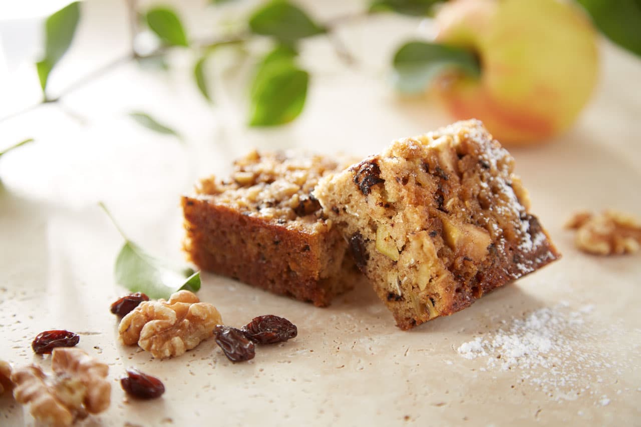 Two pieces of nut and fruit cake sit on a light surface, surrounded by walnuts, raisins, powdered sugar, and a blurred apple and leaves in the background.