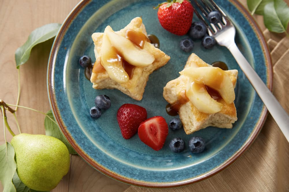 Blue plate with two star-shaped shortcakes topped with sliced pears and sauce, surrounded by blueberries and strawberries, with a fork on the plate. Pear and leaves on the table.