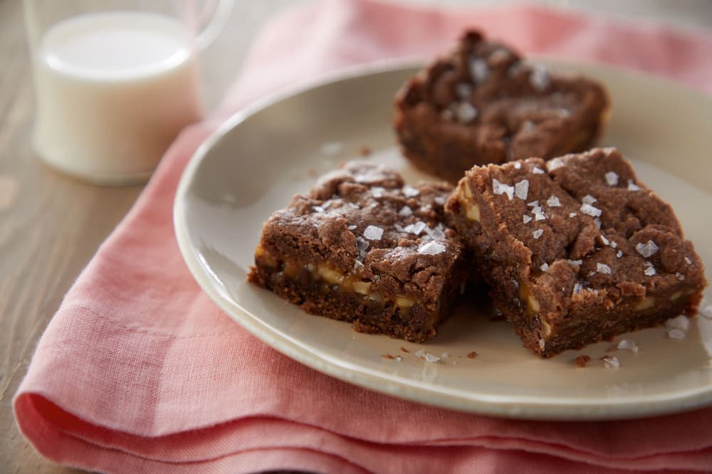 Three chocolate brownies sprinkled with flaky salt are arranged on a beige plate, set on a pink cloth with a glass of milk in the background.