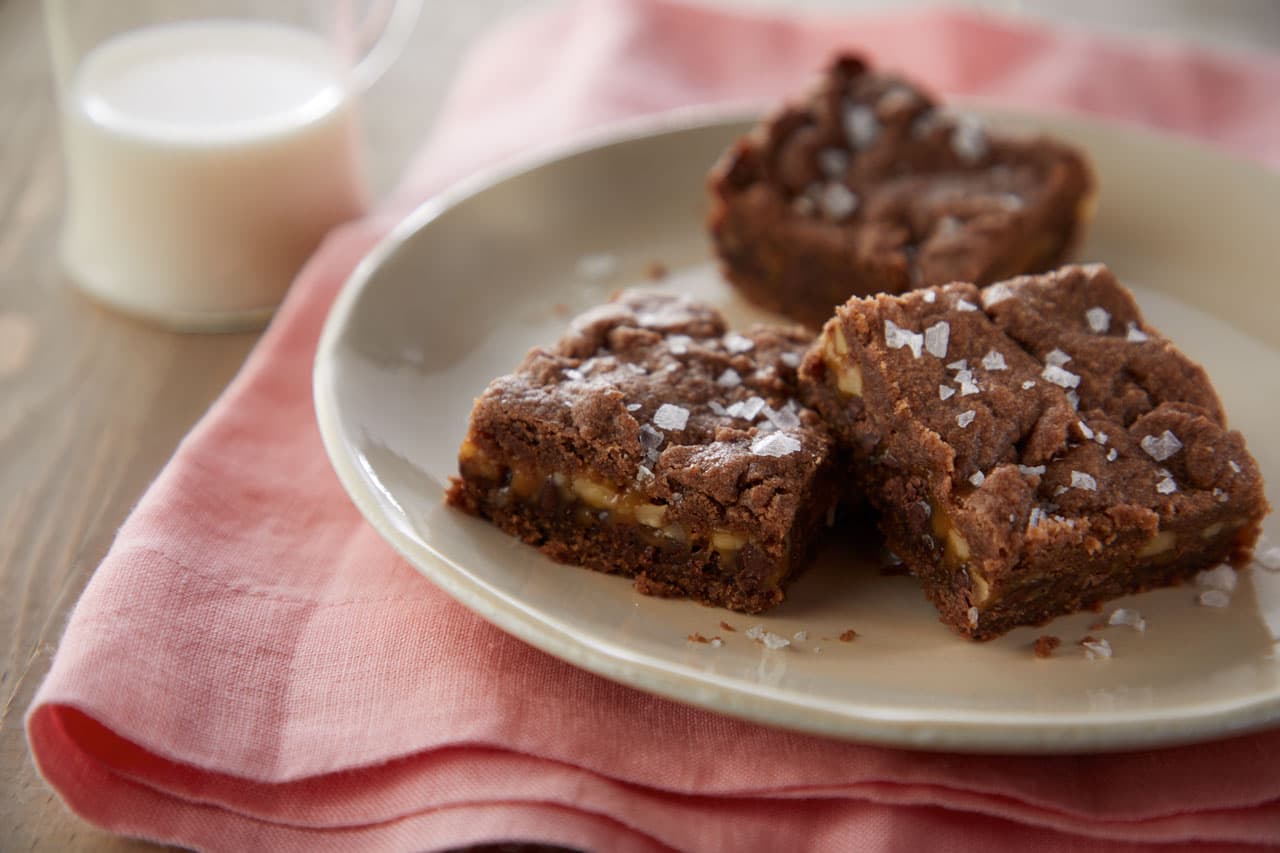 Three chocolate brownies sprinkled with flaky salt are arranged on a beige plate, set on a pink cloth with a glass of milk in the background.