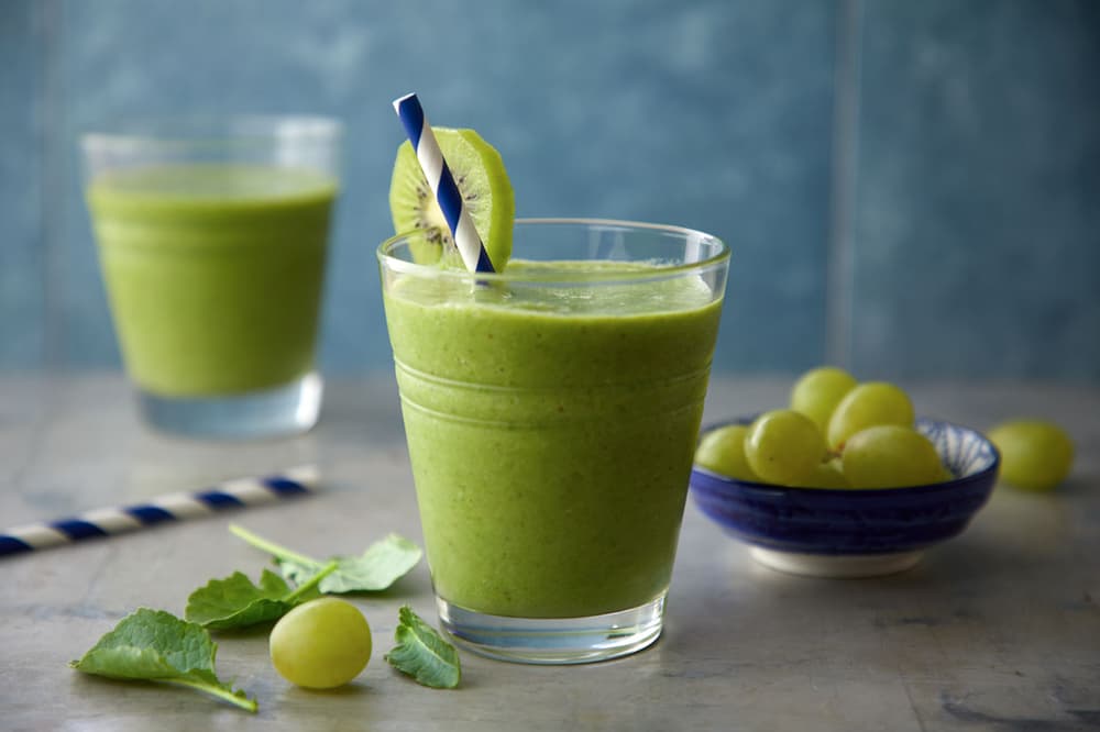 A glass of green smoothie with a kiwi slice and striped straw, next to a bowl of green grapes and leafy greens. Another smoothie glass is in the background.