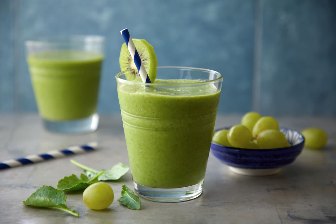 A glass of green smoothie with a kiwi slice and striped straw, next to a bowl of green grapes and leafy greens. Another smoothie glass is in the background.