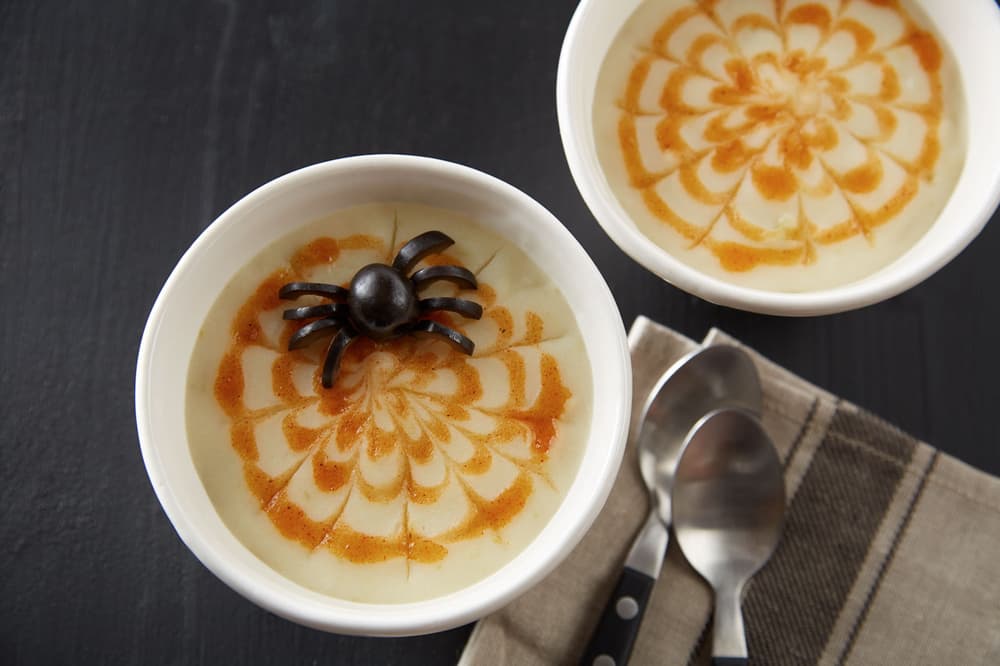 Two bowls of creamy dessert with caramel swirl designs; one bowl has a black olive spider decoration on top. Two metal spoons rest on a striped napkin beside the bowls.