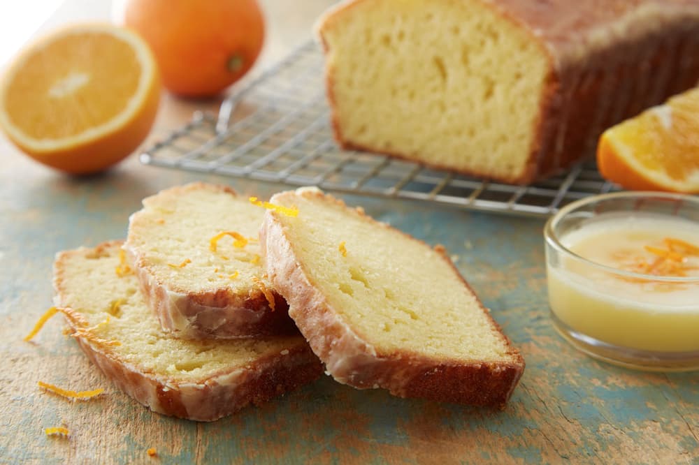 Sliced glazed orange loaf cake on a table, with fresh orange halves, a small bowl of glaze, and a cooling rack holding the rest of the loaf in the background.