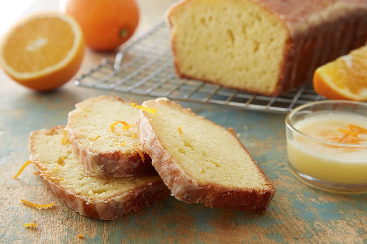 Sliced glazed orange loaf cake on a table, with fresh orange halves, a small bowl of glaze, and a cooling rack holding the rest of the loaf in the background.