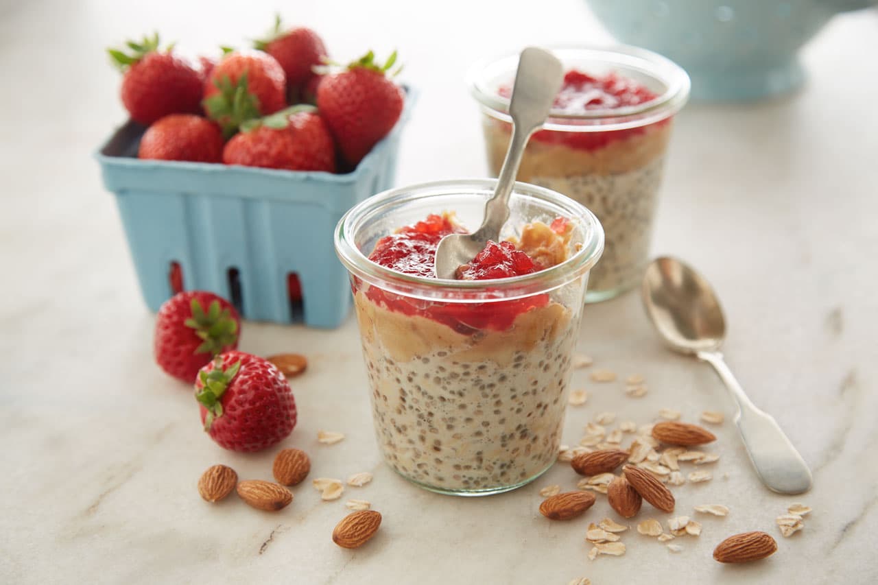 Glass jars filled with overnight oats, chia seeds, and strawberry jam, with fresh strawberries, almonds, and a blue carton of berries on a marble surface. Two metal spoons are beside the jars.