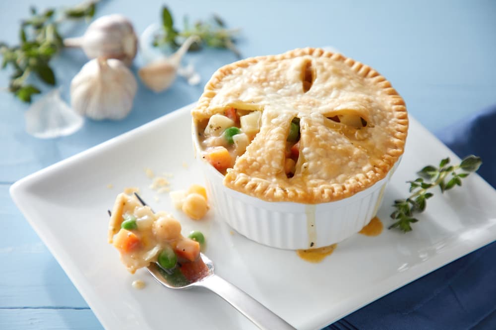 A baked pot pie in a white ramekin with a golden crust, filled with vegetables and sauce, sits on a white plate with a forkful beside it. Garlic and herbs are in the background.