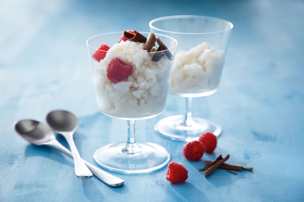 Two glass dessert cups filled with white granita, garnished with raspberries and cinnamon sticks, sit on a blue surface beside three spoons and extra garnishes.