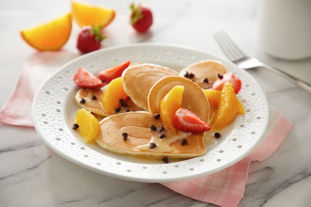A white plate with mini pancakes, orange slices, strawberries, chocolate chips, and a drizzle of cream, set on a pink napkin with a fork in the background.