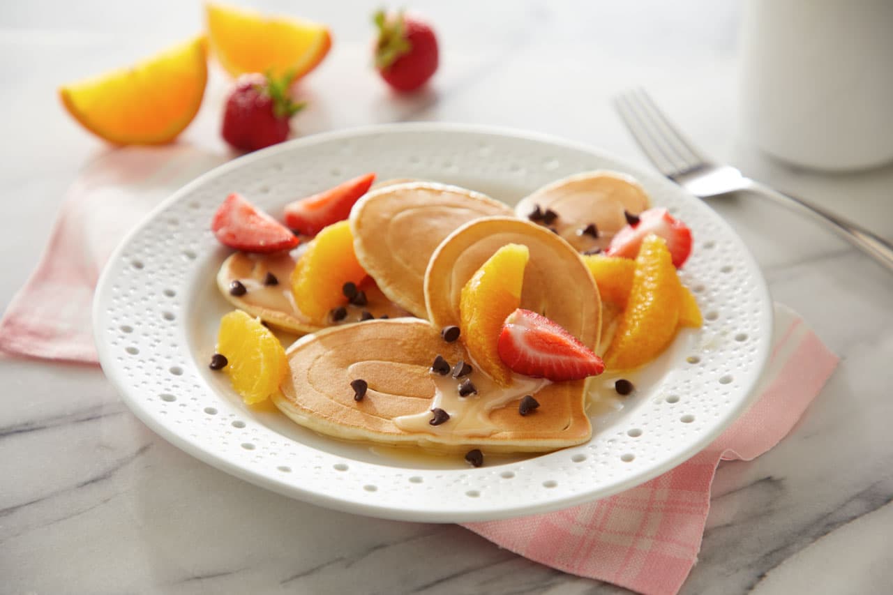 A white plate with mini pancakes, orange slices, strawberries, chocolate chips, and a drizzle of cream, set on a pink napkin with a fork in the background.