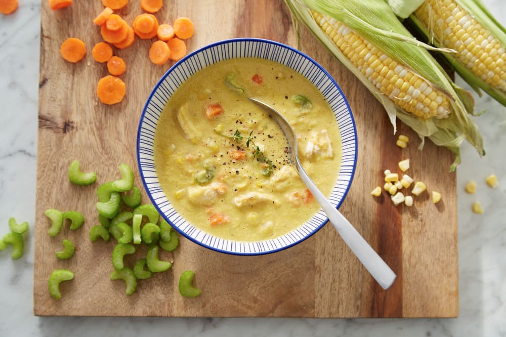 A bowl of corn chowder with a spoon sits on a cutting board beside raw celery, sliced carrots, and fresh corn on the cob.
