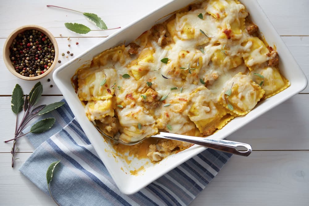 Baked ravioli casserole topped with melted cheese and herbs in a white dish, with a serving spoon, striped towel, peppercorns, and sage leaves on a white wooden table.