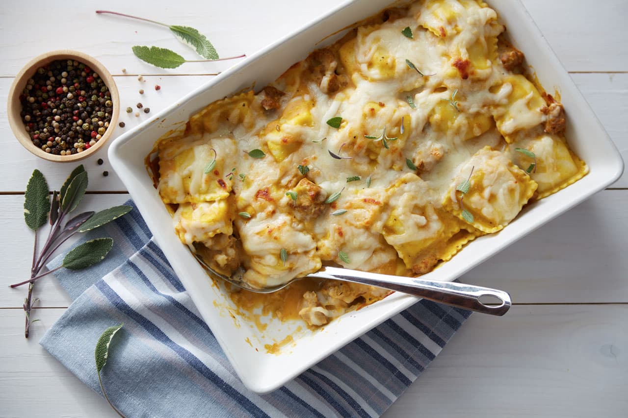 Baked ravioli casserole topped with melted cheese and herbs in a white dish, with a serving spoon, striped towel, peppercorns, and sage leaves on a white wooden table.