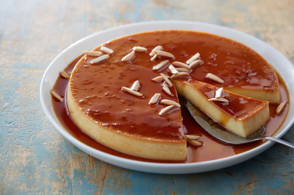 Round flan with caramel sauce on a white plate, topped with slivered almonds. A slice is cut and lifted on a metal spatula.