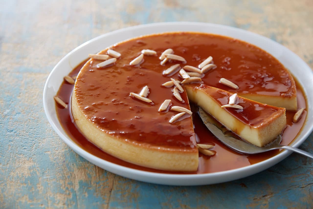 Round flan with caramel sauce on a white plate, topped with slivered almonds. A slice is cut and lifted on a metal spatula.