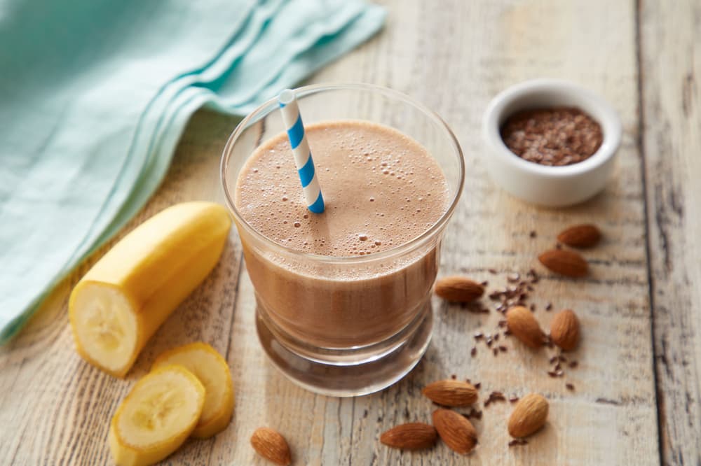 A glass of chocolate smoothie with a striped straw sits on a wooden table, surrounded by banana slices, almonds, flaxseeds, and a napkin.