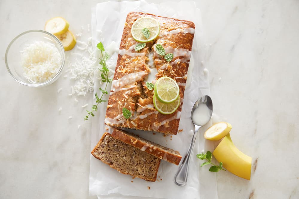 A loaf of banana bread with icing and lime slices on top, a slice cut, surrounded by banana pieces, shredded coconut, mint, and a spoon on a white surface.