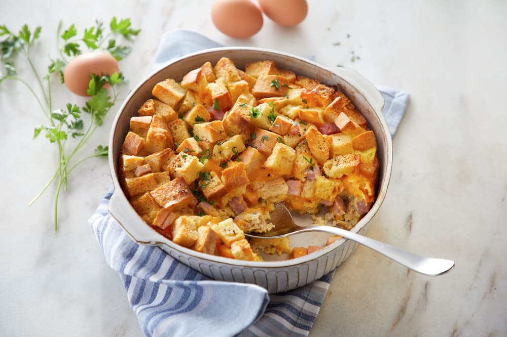 A casserole with cubed bread, ham, and cheese sits in a round baking dish with a spoon. Fresh parsley and eggs are on the white marble surface beside the dish.