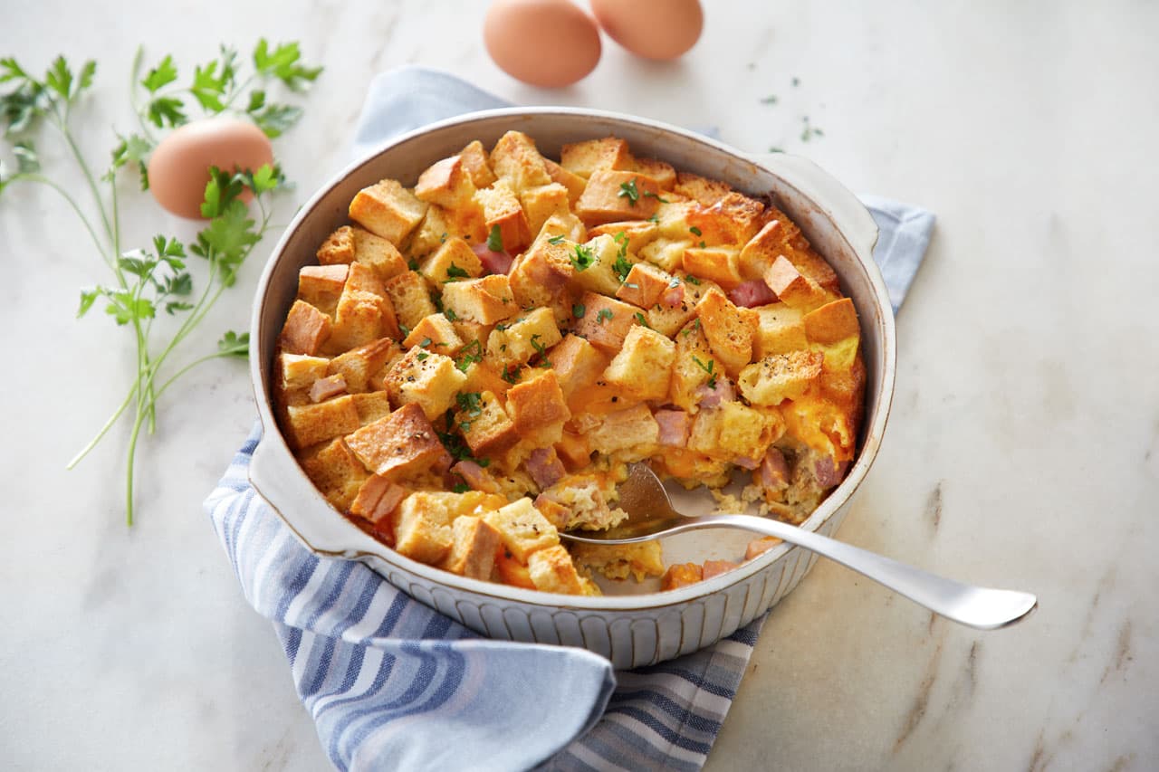 A casserole with cubed bread, ham, and cheese sits in a round baking dish with a spoon. Fresh parsley and eggs are on the white marble surface beside the dish.