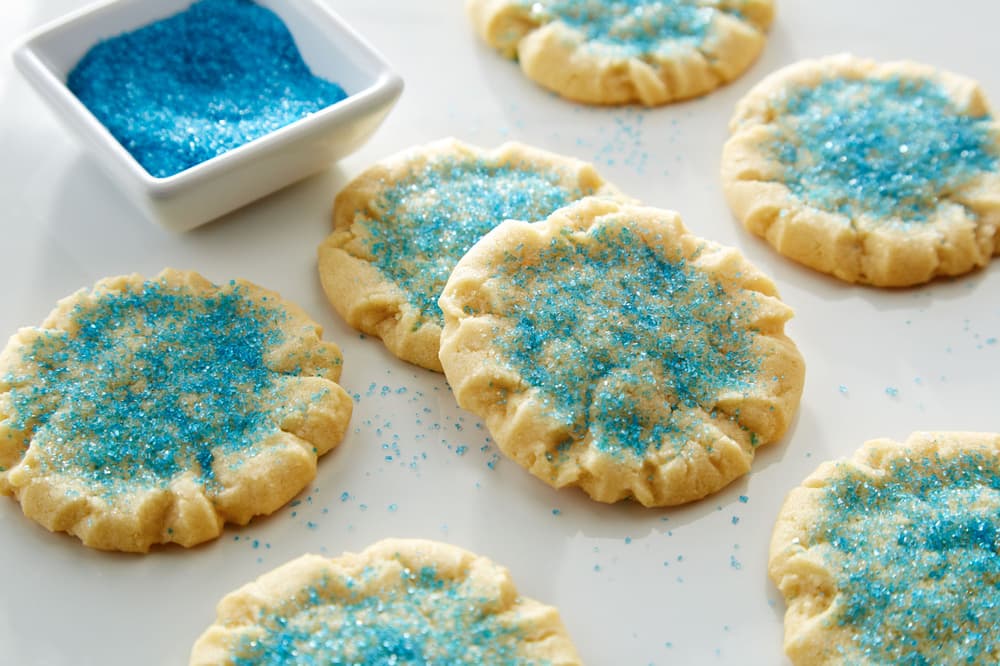 Sugar cookies topped with blue sugar sprinkles are arranged on a white surface, with a small dish of extra blue sprinkles nearby.