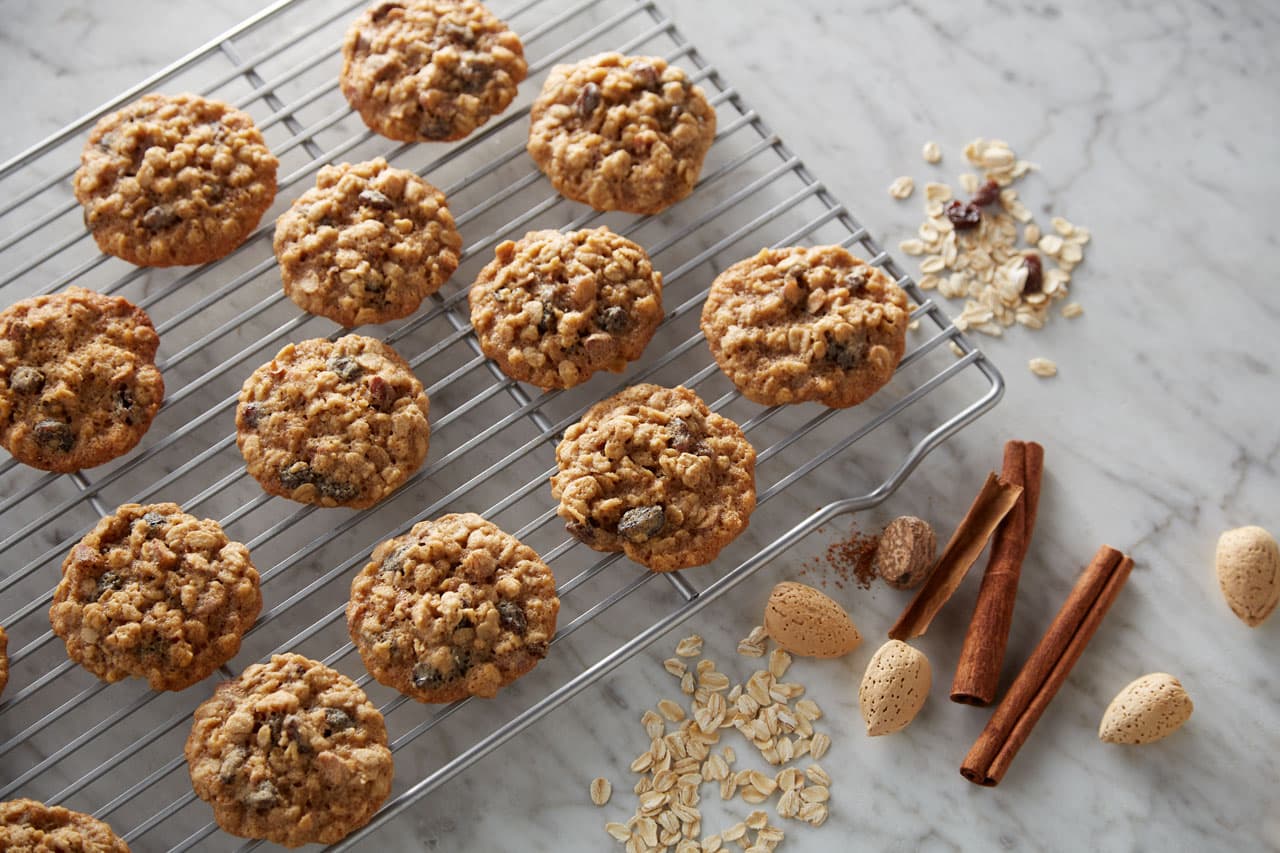 Oatmeal cookies cooling on a metal rack, with oats, whole almonds, cinnamon sticks, and raisins scattered on a marble surface nearby.