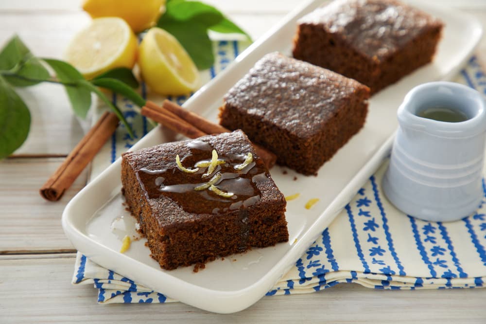 Three pieces of brown cake on a white tray, garnished with lemon zest, with a small pitcher, cinnamon sticks, and lemons in the background.