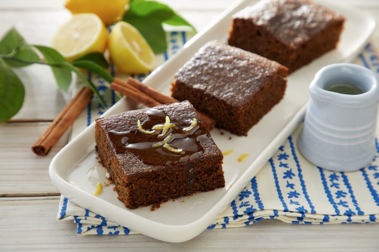Three pieces of brown cake on a white tray, garnished with lemon zest, with a small pitcher, cinnamon sticks, and lemons in the background.