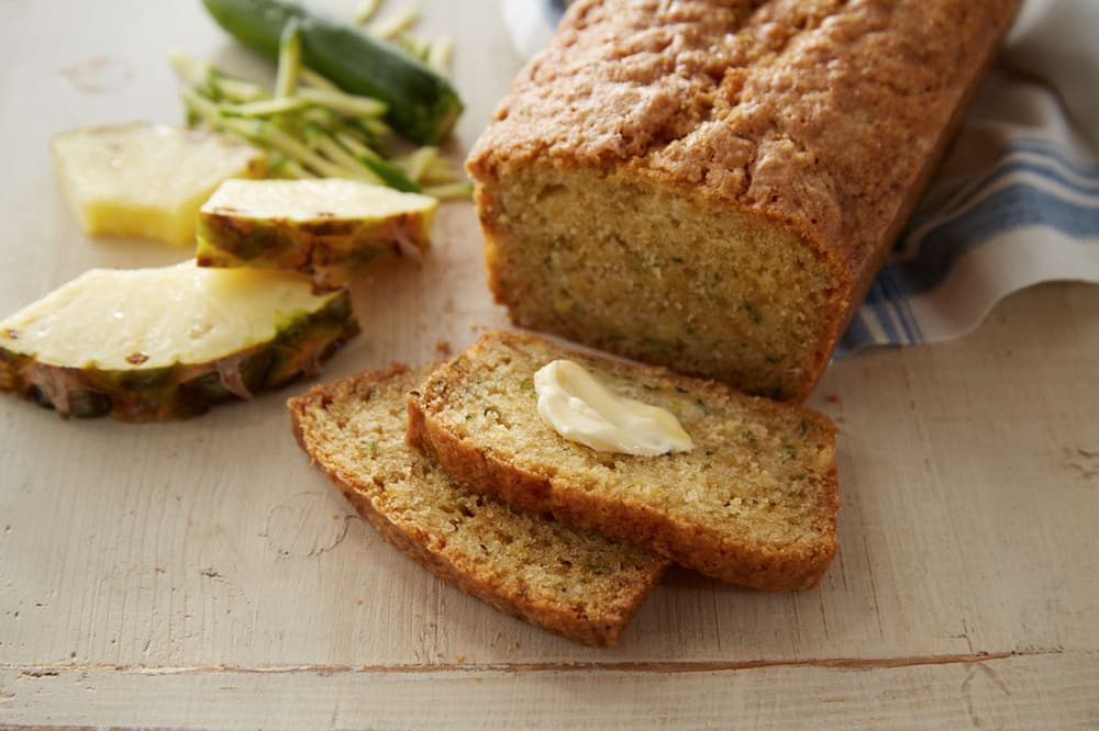 Sliced zucchini bread loaf with a pat of butter on one slice, next to pineapple slices and shredded zucchini on a wooden surface.
