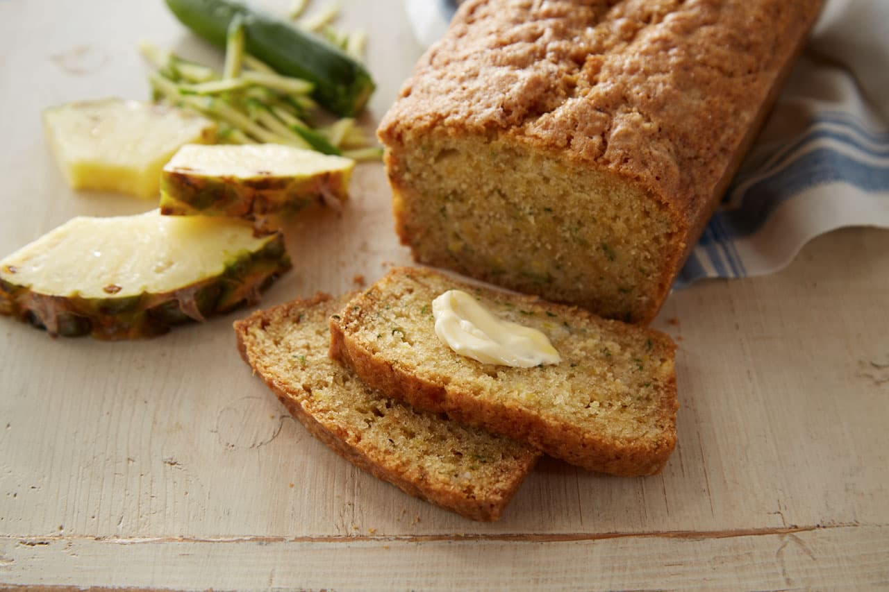 Sliced zucchini bread loaf with a pat of butter on one slice, next to pineapple slices and shredded zucchini on a wooden surface.