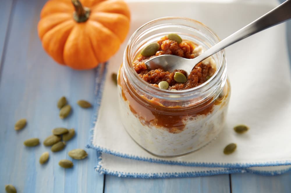 A glass jar of oatmeal topped with pumpkin puree and seeds sits on a napkin, with a spoon inside. Pumpkin and pumpkin seeds are placed nearby on a blue wooden surface.