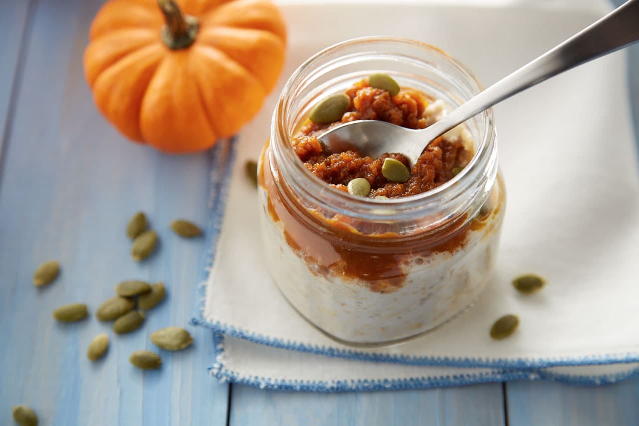 A glass jar of oatmeal topped with pumpkin puree and seeds sits on a napkin, with a spoon inside. Pumpkin and pumpkin seeds are placed nearby on a blue wooden surface.
