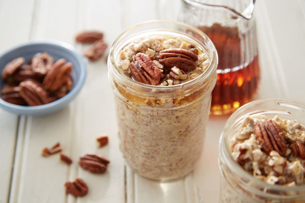 A mason jar filled with overnight oats topped with pecans, next to a plate of pecans and a glass pitcher of syrup on a white surface.