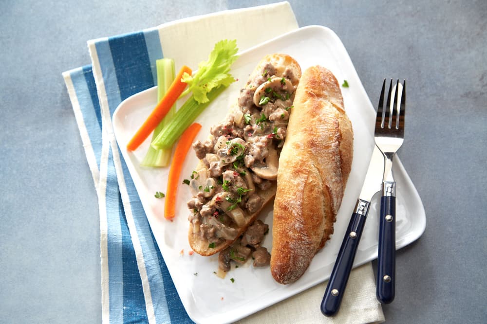 Open-faced sandwich with creamy beef and mushroom topping, served with a baguette, celery, carrot sticks, and utensils on a white plate.
