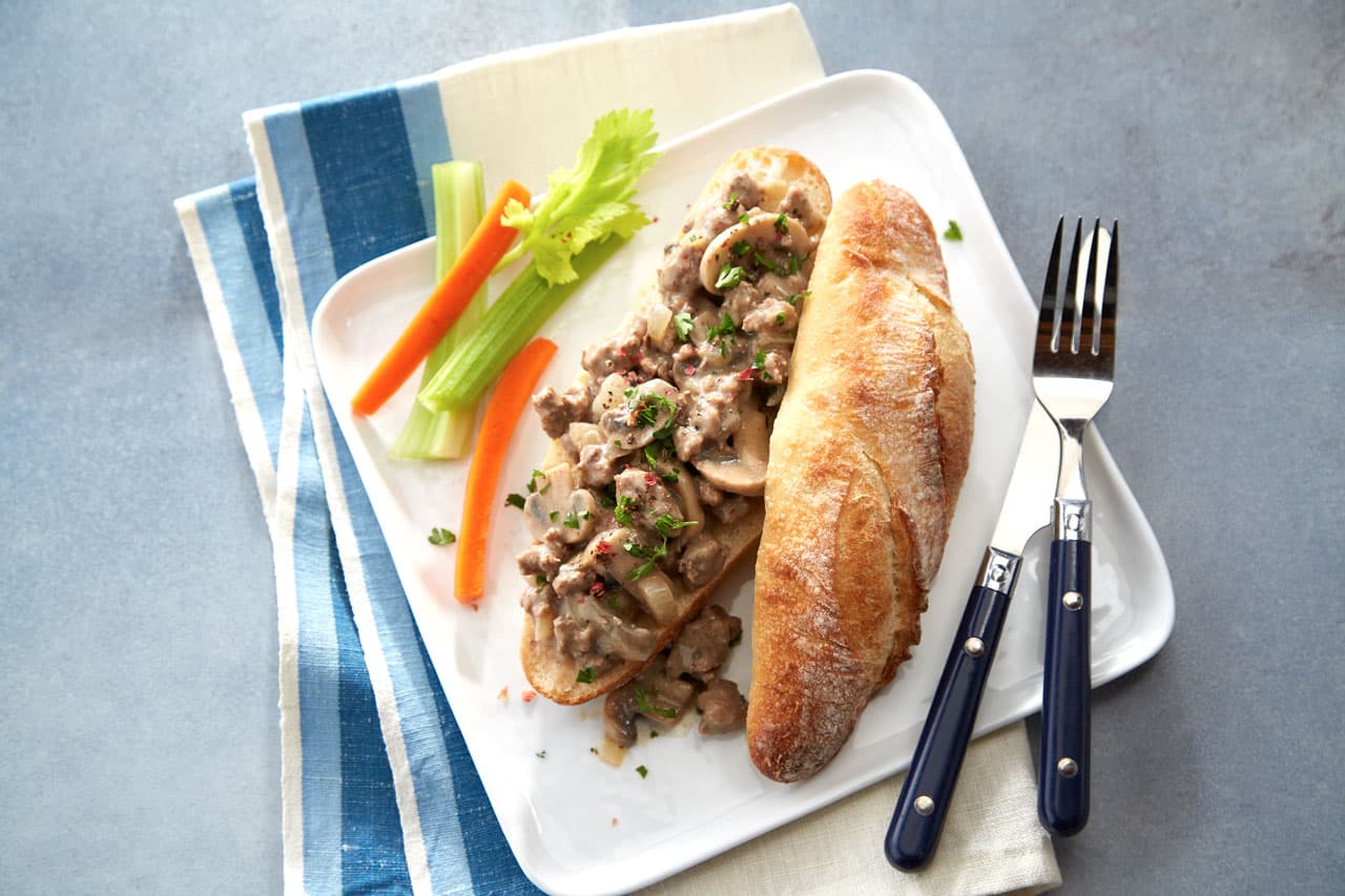 Open-faced sandwich with creamy beef and mushroom topping, served with a baguette, celery, carrot sticks, and utensils on a white plate.