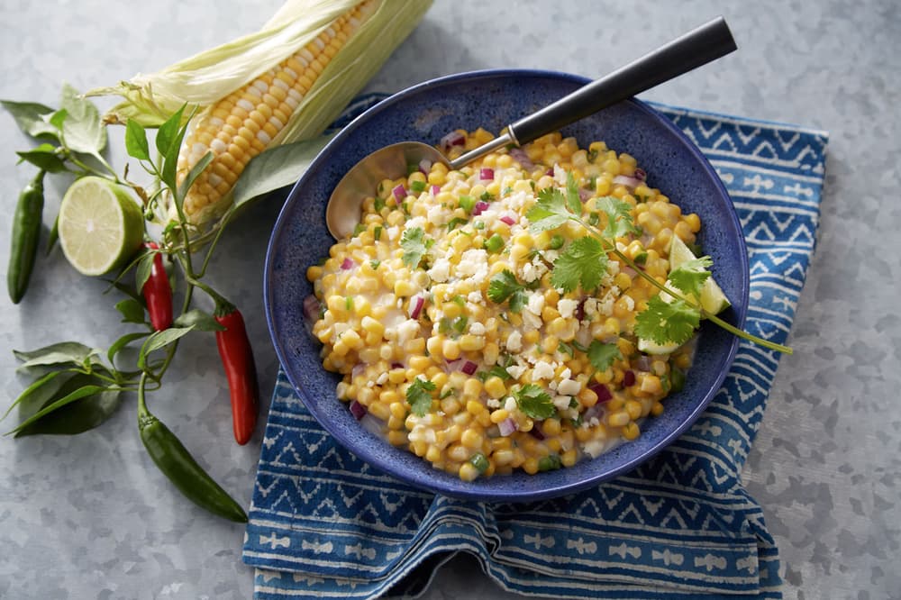 A blue bowl of corn salad with red onion, cilantro, and cheese sits on a patterned cloth, next to fresh corn, lime, and chili peppers.