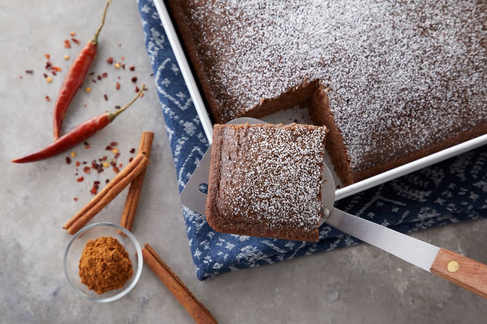 A square piece of chocolate cake with powdered sugar is lifted from a tray; cinnamon sticks, chili peppers, and ground spice are nearby.