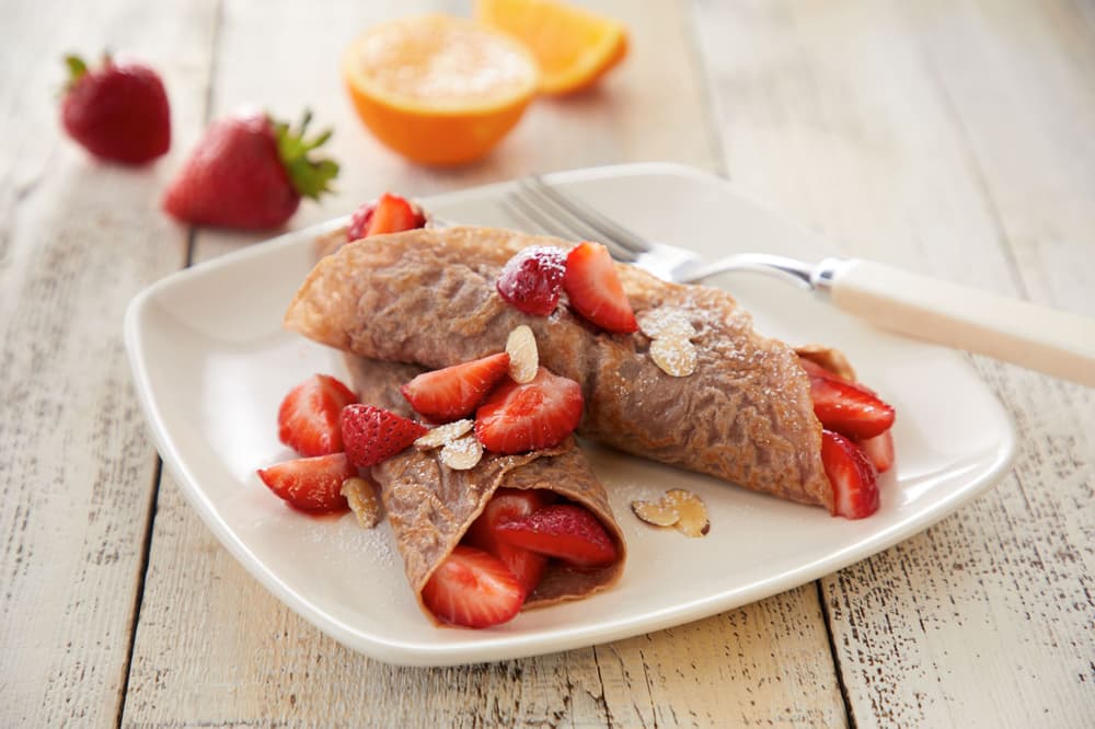 Two chocolate crepes filled with sliced strawberries on a white plate, garnished with oats and powdered sugar, with a fork and fruit in the background.