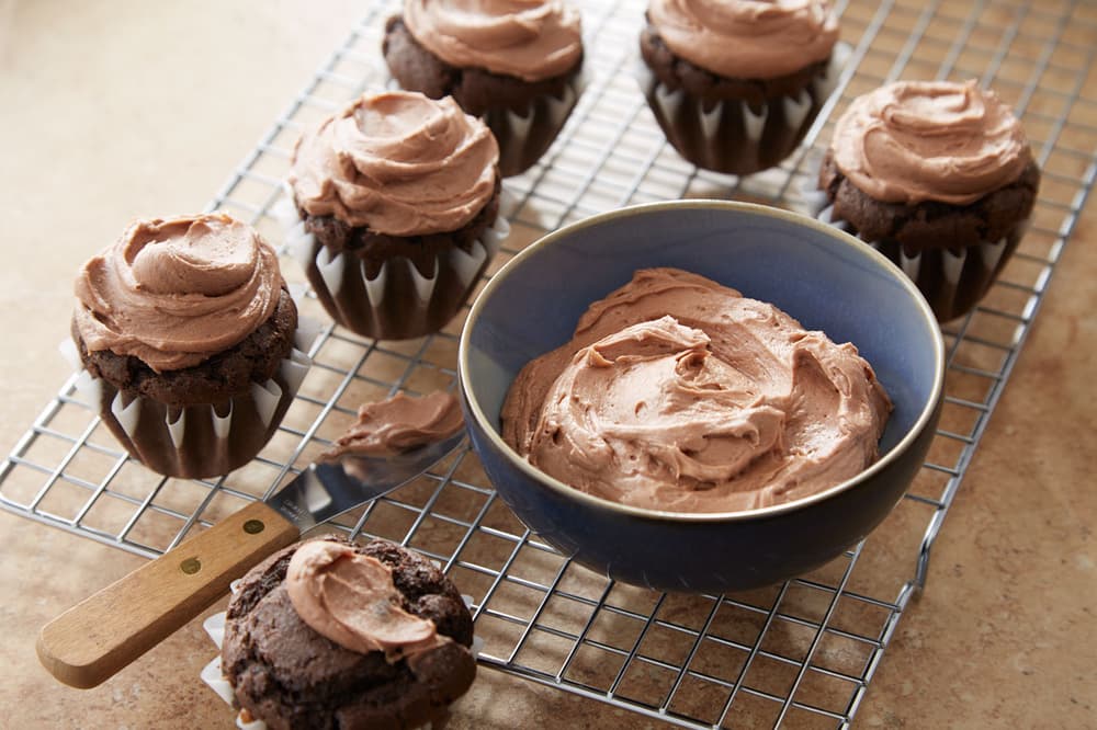 Chocolate cupcakes with chocolate frosting are placed on a wire rack, with a bowl of extra frosting and a butter knife beside them.