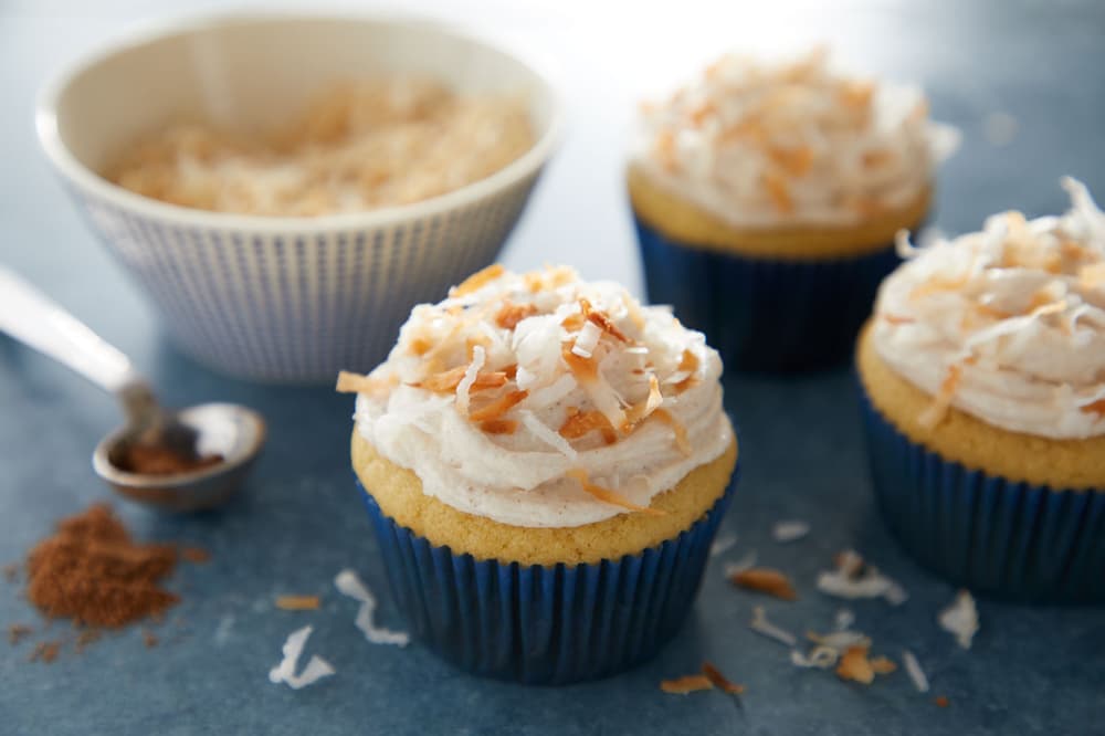Three cupcakes with white frosting and toasted coconut topping are arranged on a blue surface, with a bowl of shredded coconut and a spoon with spice in the background.